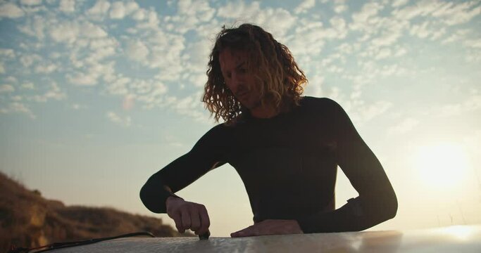 Close-up shot of a young guy with curly hair in a black wetsuit cleaning a surfboard using a special tool against the background of a blue sky and white clouds in the sun in the morning in summer