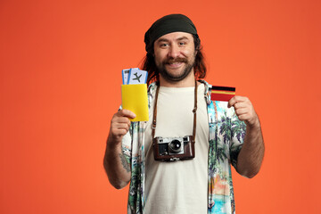 Happy male traveler holding passport with airline tickets and credit card. Man with camera around his neck in studio on orange background close up. - obrazy, fototapety, plakaty