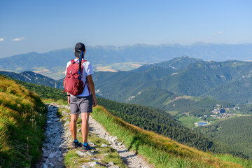 Polish Tatra Mountains, a young man in shorts and a cap with a mountain backpack stands on a mountain trail and looks at the valley with mountain peaks on a summer sunny day. - obrazy, fototapety, plakaty