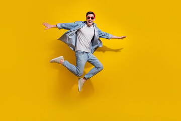 Full length photo of excited cheerful guy wear denim jacket dark glasses jumping high isolated yellow color background - obrazy, fototapety, plakaty