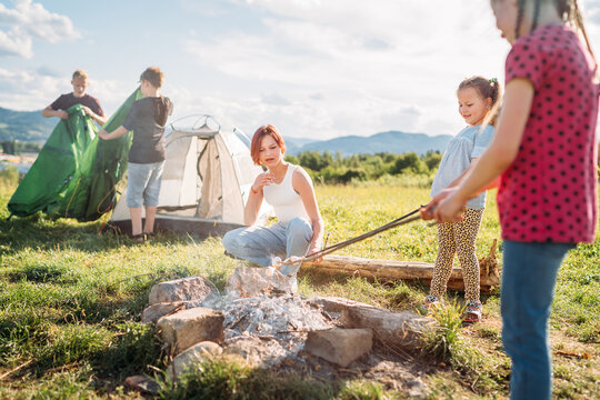 Three sisters have picnic, roasting marshmallows and candies on sticks over campfire flame while two brothers set up the green tent. Happy family outdoor picnic camping activities concept