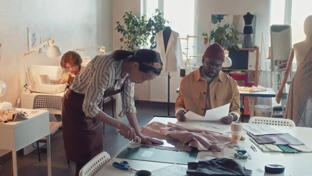 Side shot of diverse skilled atelier workers during garment production process preparing blanks and drawing sketches while creating new clothing collection in modern tailoring studio