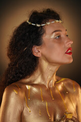 Portrait of female model in studio. Close up shot of attractive girl wearing tiara and red lipstick, body covered in golden paint looking away. © kinomaster
