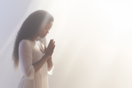 Pretty young African american woman praying to god with her eyes closed and clasped hands - profile side angle - God's rays of light shining down - Ethnic diversity and religion concept
