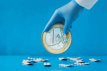 A hand in a blue medical glove holds a large 1 euro coin over scattered white tablets in blister packs on a blue background. Money for medicine, money for drugs, money for expensive treatment - obrazy, fototapety, plakaty