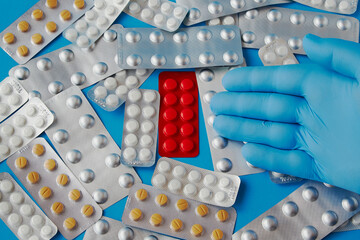 A hand in a blue medical glove points to a blister of red pills among scattered different pills in...