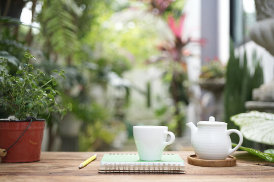 White tea kettle and white cup with notebook on wooden table outdoor garden view