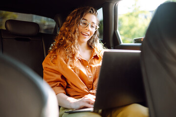 Beautiful business woman is using a laptop while sitting in the back seat of a car. Young freelancer woman working on a laptop in a car. Freelance, transportation concept.