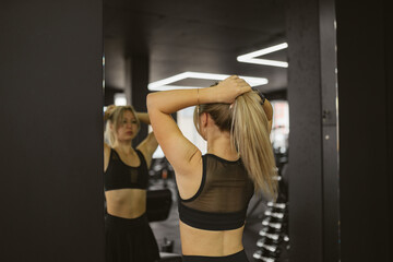 Focused woman tying hair in ponytail looking in mirror in sports club with modern fitness...