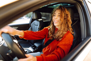 Young smiling woman driving a car drives around the city. Curly-haired woman travels by car. Lifestyle concept. - obrazy, fototapety, plakaty