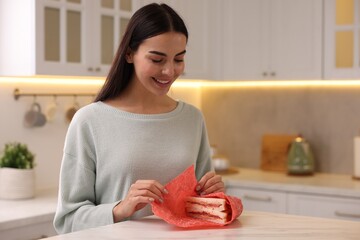 Happy woman packing sandwich into beeswax food wrap at table in kitchen - obrazy, fototapety, plakaty