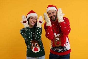 Young couple in Christmas sweaters, Santa hats and knitted mittens on orange background