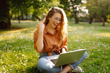 Curly-haired woman in casual clothes with a laptop sits on the lawn on a sunny day. Happy freelancer woman working outdoors and enjoying the weather.