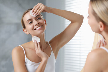 Smiling woman removing makeup with cotton pads in front of mirror indoors