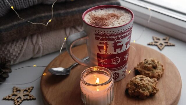 Winter windowsill still life. Red ceramic cup of hot coffee on window sill. Christmas decorations on the background. Cozy home picture. Warm woolen knitted sweaters, Burn Candle, Cookies. Stock photo