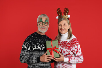 Senior couple in Christmas sweaters, reindeer headband and funny glasses holding gift on red...