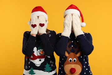 Couple in Christmas sweaters and Santa hats covering faces with hands in knitted mittens on orange background