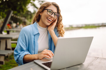 Portrait of a young woman in a blue shirt with a laptop at a table in an outdoor cafe near a lake. Curly woman freelancer works on a laptop outdoors. Freelance concept, nature.