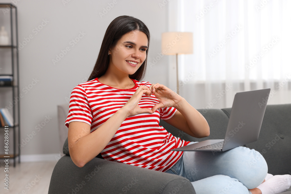 Wall mural Happy young woman having video chat via laptop and making heart with hands on sofa in living room