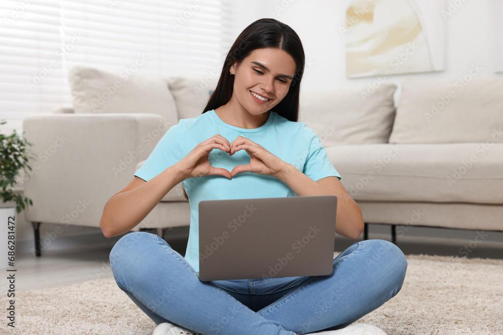 Wall mural Happy young woman having video chat via laptop and making heart on floor in living room