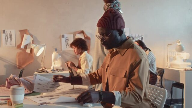 Medium side shot of African American male stylish fashion designer reworking multiple sketches of new apparel collection during workday in atelier