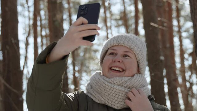 Joyful white european woman takes selfie on her phone in frosty winter forest. Video call via mobile Internet while traveling on sunny day. Happy influencer in warm clothes shoots video in nature.