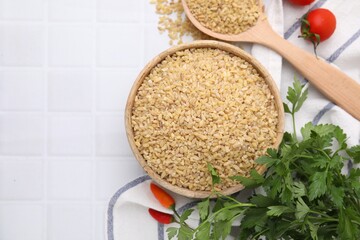 Raw bulgur in bowl, spoon, vegetables and parsley on white tiled table, flat lay. Space for text