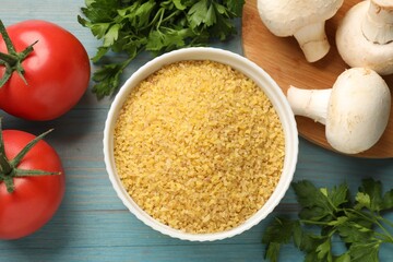 Raw bulgur in bowl, vegetables and parsley on light blue wooden table, flat lay