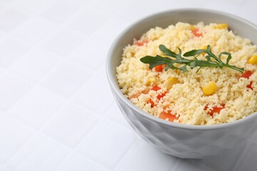 Tasty couscous with pepper, corn and arugula in bowl on white tiled table, closeup. Space for text