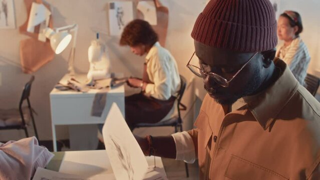 Side tilt shot of Black male clothes designer in glasses and beanie looking through multiple finished sketches during workday in atelier