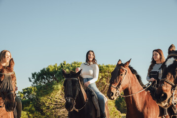 Cheerful girlfriends riding horses against evergreen tree and blue sky
