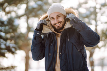A young bearded man walks in a snowy park. Happy man enjoying the weather on a winter sunny day. Concept of walks, weekends. - obrazy, fototapety, plakaty