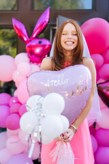 Obraz na płótnie Canvas Vertical portrait of cheerful redhead birthday female in summer dress posing holding various colorful festive foil helium balloons standing by country house, closed eyes with happy expression.