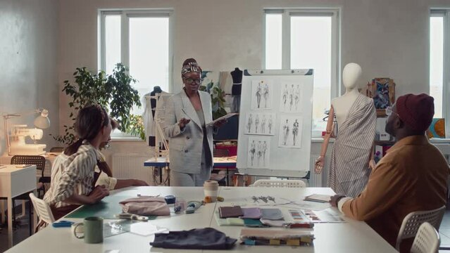 Full shot of diverse atelier workers sitting at table and business owner coming with tablet and speaking about new apparel collection using whiteboard with sketches in tailoring studio