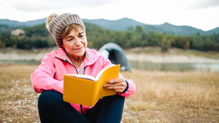 Happy senior woman reading a book outdoors during hiking day. Adventure and travel vacations concept - obrazy, fototapety, plakaty
