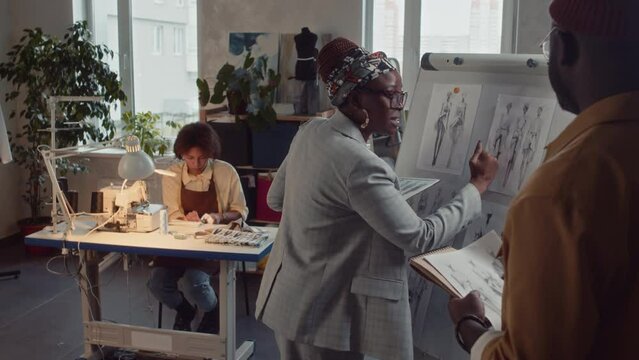 Medium side shot of female creative director discussing ideas for new clothing collection with unrecognizable male designer using sketches on whiteboard and tablet at atelier