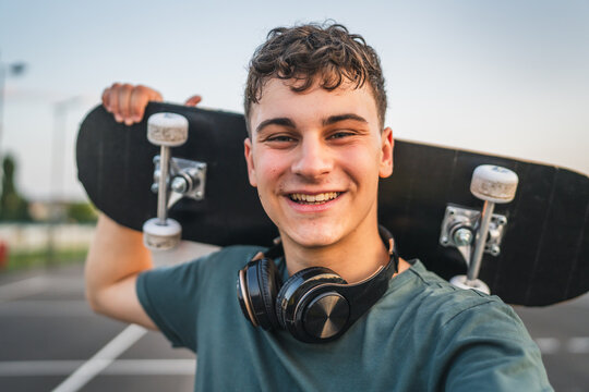 One man young adult caucasian teenager stand outdoor with skateboard on his shoulder and headphones posing at basketball court portrait happy confident wear shirt casual real person copy space