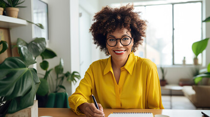 Woman with curly hair and glasses is writing in a notebook at a home office - obrazy, fototapety, plakaty