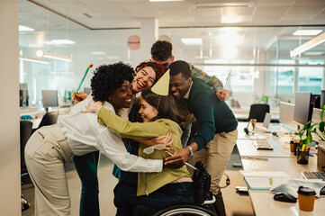 Diverse colleagues hugging birthday girl who uses a wheelchair at the office.