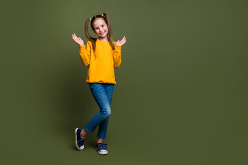 Photo of cute cheerful schoolgirl with ponytails wear yellow pullover stand near empty space palms up isolated on khaki color background