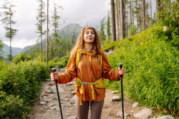 Young female traveler with hiking poles and a bright backpack enjoys the mountain scenery along a...