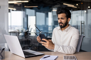 Young Indian man businessman working in office, worriedly holding glasses in hand and looking at mobile phone