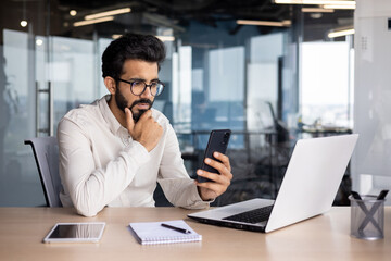 Pensive young Indian man businessman sits in the office at the table, looks at the phone, reads...