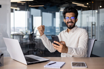 Fototapeta na wymiar Portrait of a young Indian businessman sitting in the office at the table, holding the phone in his hands and looking at the camera, showing a victory gesture with his hand