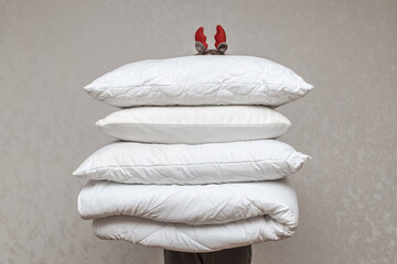 Man in santa hat holding a warm duvet and feather pillows against a gray wall. Stack of bedding for...