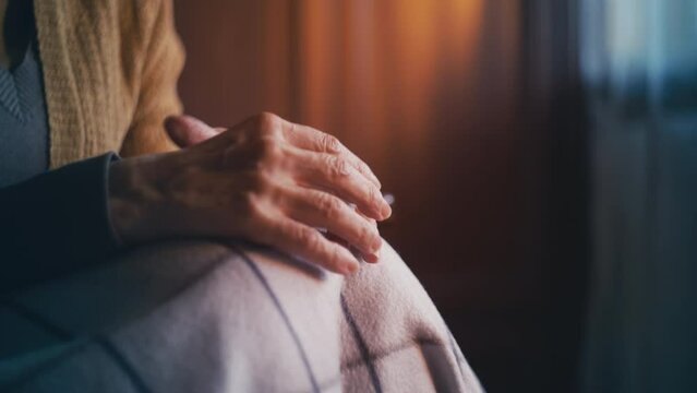 Senior woman covered in blanket sitting in armchair, close-up of trembling hands