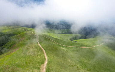 Aerial drone flight through the clouds, above an alpine mountain pasture crossed by a dirt road....