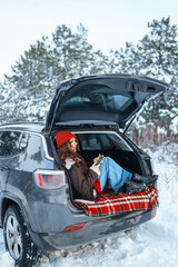 Happy woman in a red hat with a phone in her hand sits in the trunk of a car in a sunny winter forest. Concept of technology, adventure, travel by car.