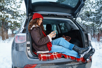 Happy woman in a red hat with a phone in her hand sits in the trunk of a car in a sunny winter forest. Concept of technology, adventure, travel by car.