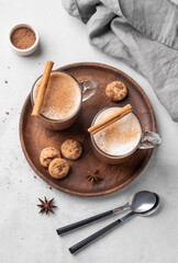 Fototapeta na wymiar Two glass mugs with hot chocolate and milk foam on a wooden board on a gray background with cinnamon sticks, anise star, spoon and cookies.